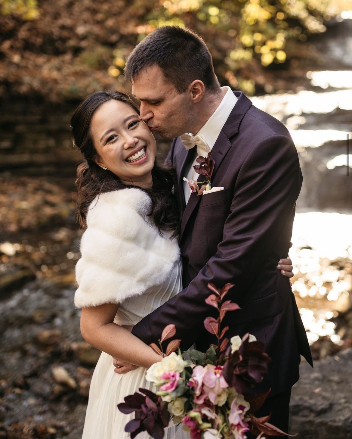 A couple posing for their wedding photos in an outdoor setting. The groom is kissing the bride on the forehead. The bride is smiling and holding a bouquet of flowers. They are situated near a stream with a backdrop of autumn foliage.