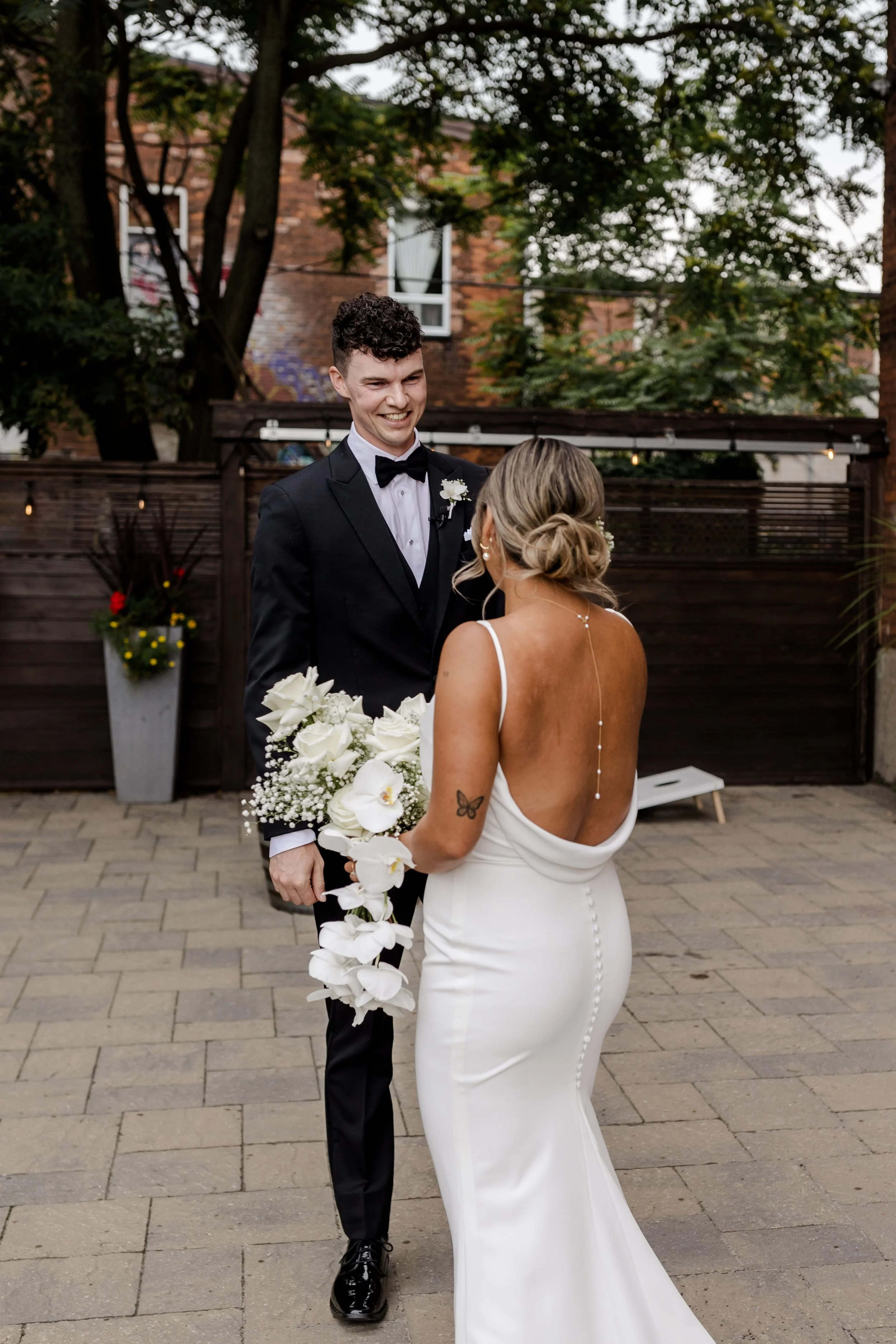 Bride and groom in elegant attire, with bride holding a bouquet of white flowers, standing outside in a garden setting.