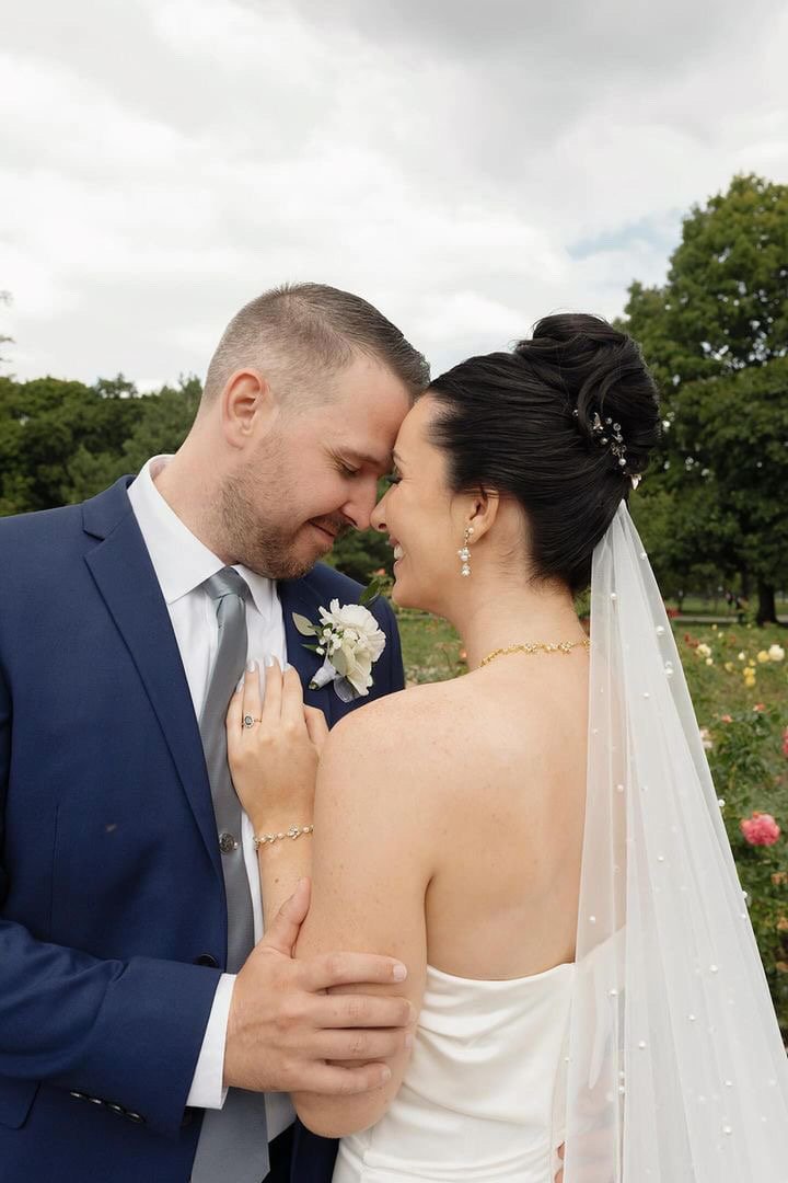 Wedding couple embracing outdoors, bride in white dress and veil, groom in blue suit and tie.