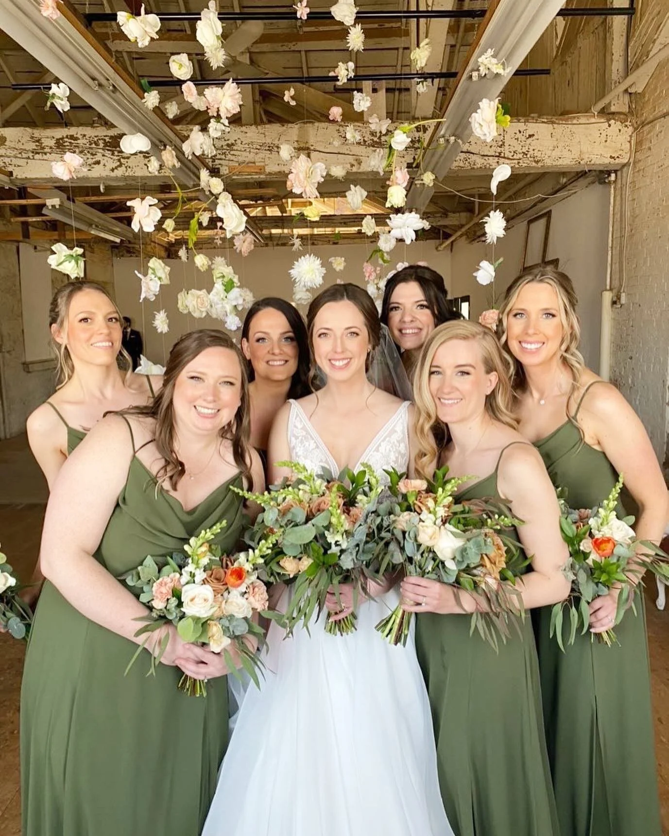 Bride with bridesmaids in green dresses holding bouquets with flowers hanging overhead.