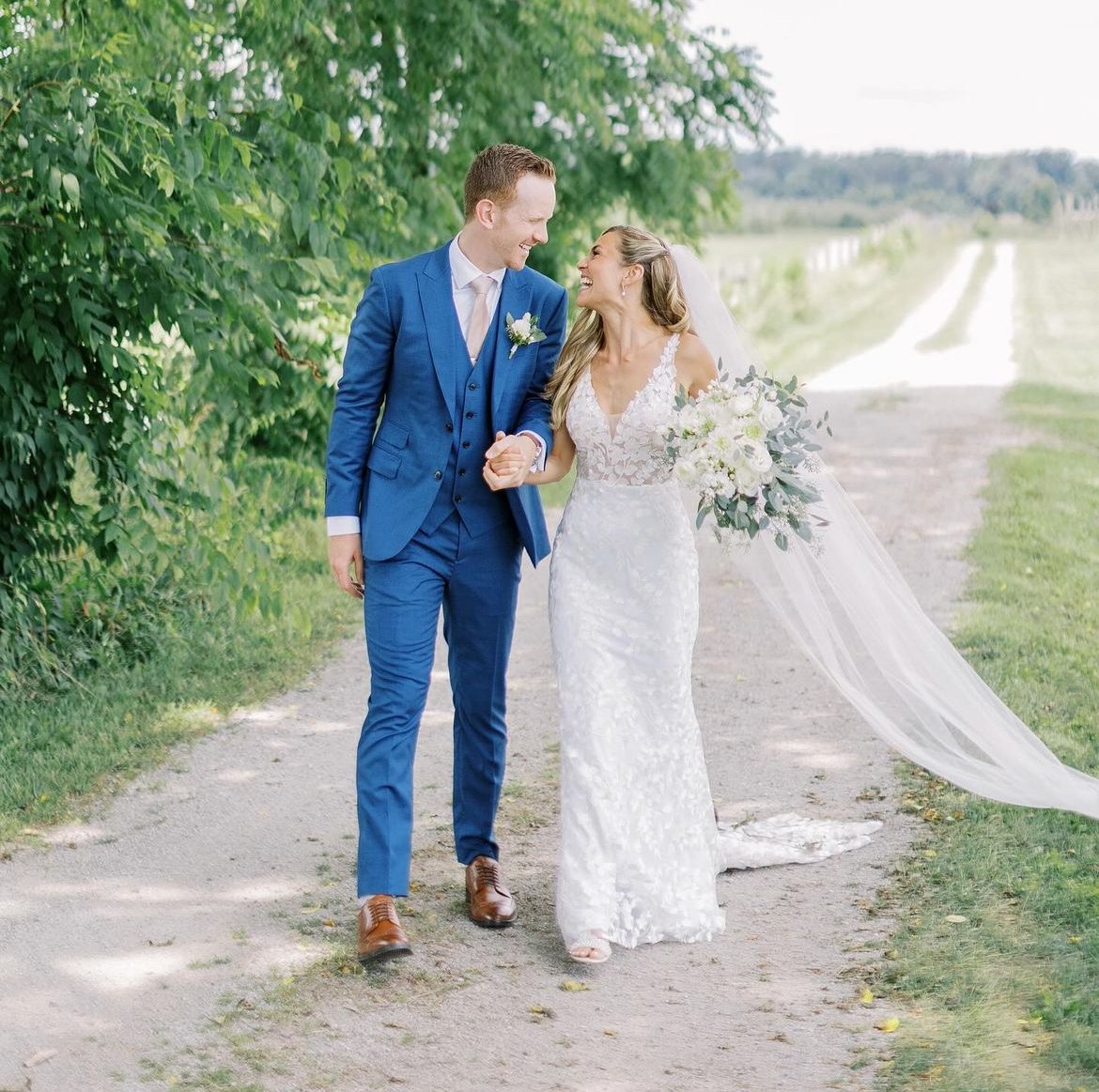 A bride in a white dress with a long veil and groom in a blue suit walking on a path, holding hands, smiling, and surrounded by greenery.