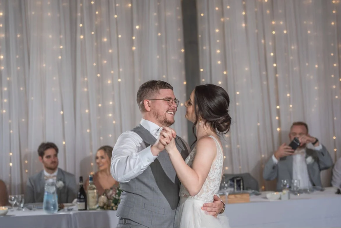 Bride and groom dancing at a wedding reception, with guests seated at a table in the background, soft lights along curtains.
