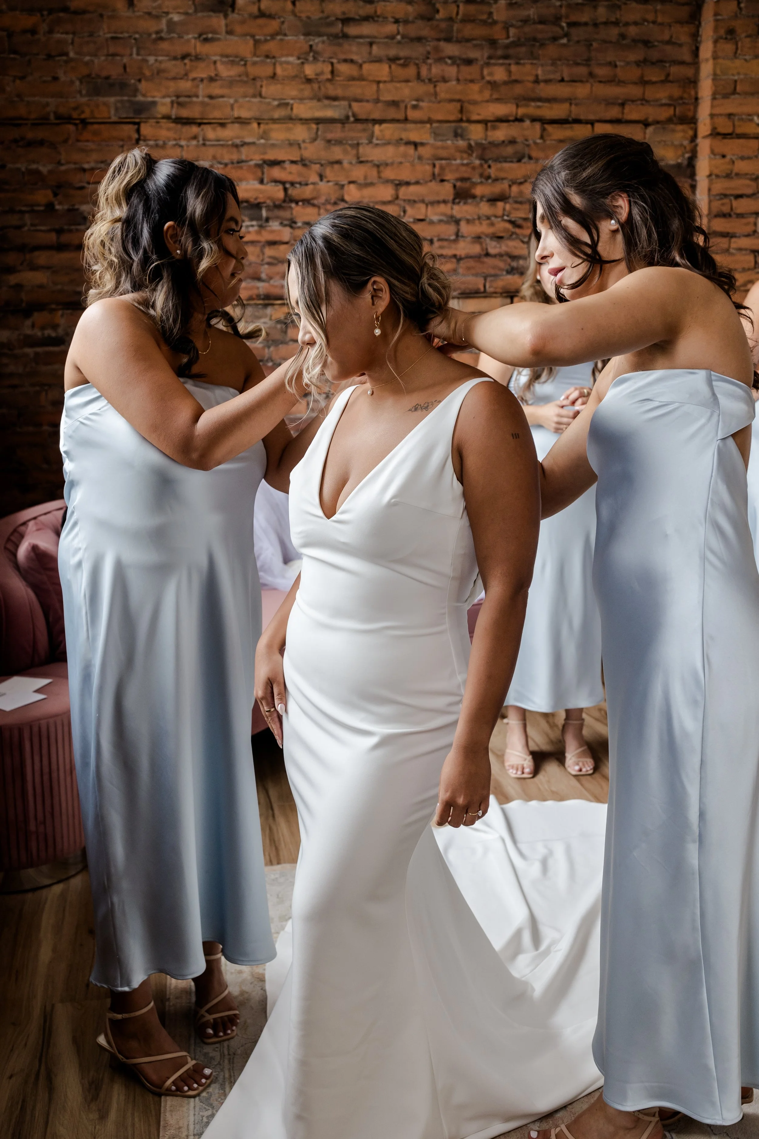 Bride in white dress being assisted by bridesmaids in blue dresses.