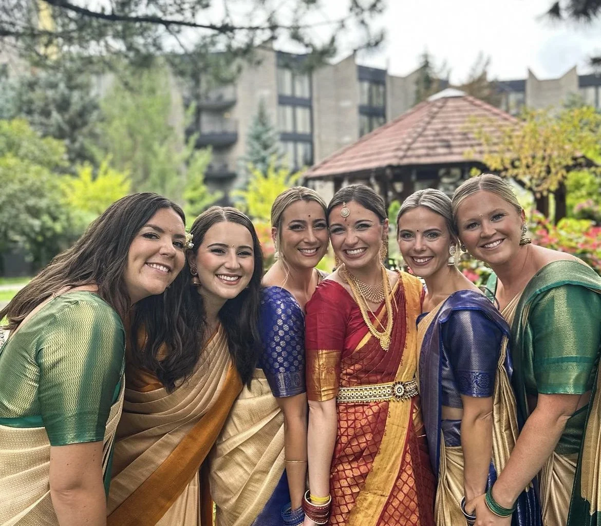 Six women standing outdoors, smiling, wearing colorful traditional Indian sarees with a garden and building in the background.