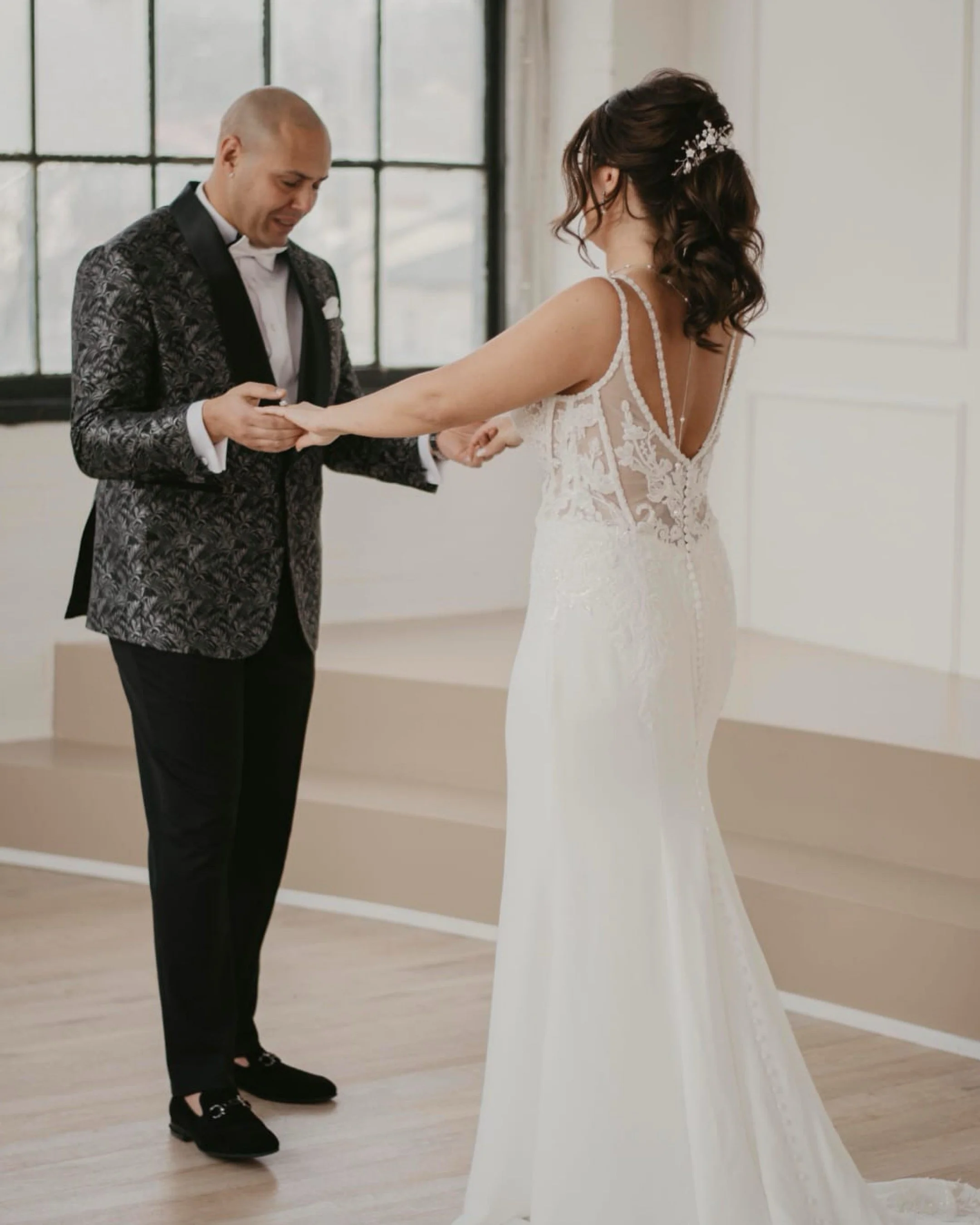 A bride in a white wedding dress and a groom in a patterned jacket holding hands in a bright room.