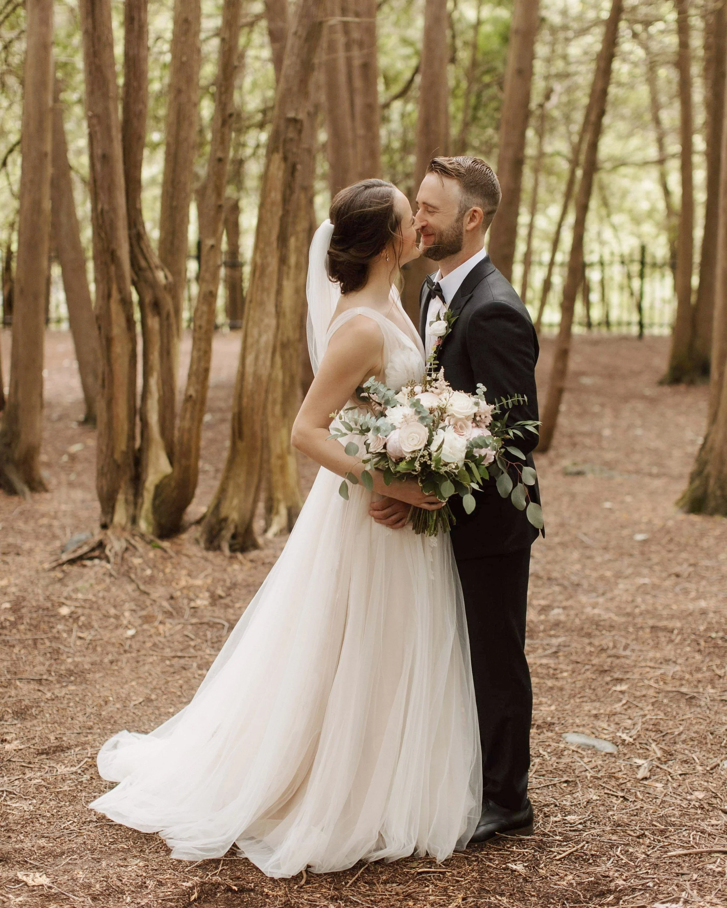 Bride and groom smiling at each other in a forest, holding a bouquet.
