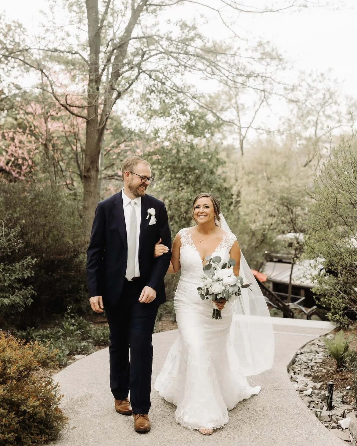 Bride and groom walking on a garden pathway, bride holding a bouquet, wearing a white lace wedding dress, groom in a navy suit, surrounded by trees.