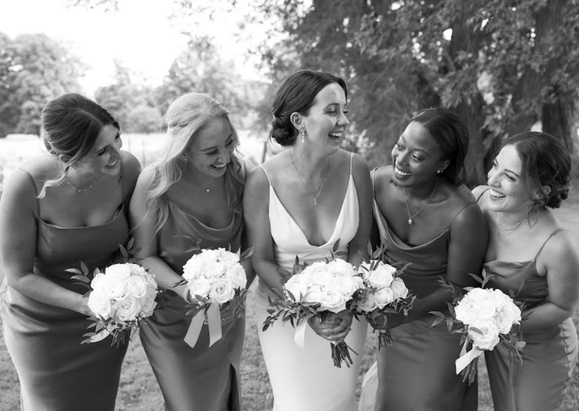 Bride and bridesmaids laughing, holding bouquets, outdoors in a garden.