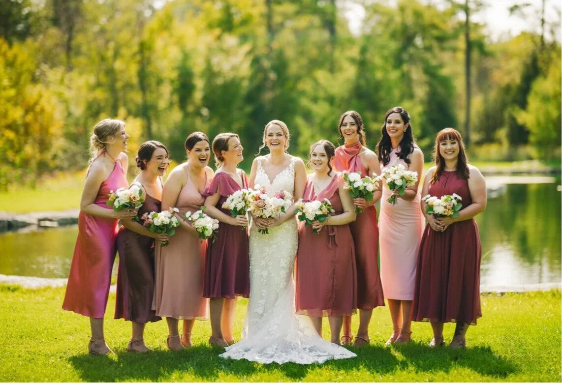 Bride and bridesmaids in colorful dresses holding bouquets outdoors by a pond.