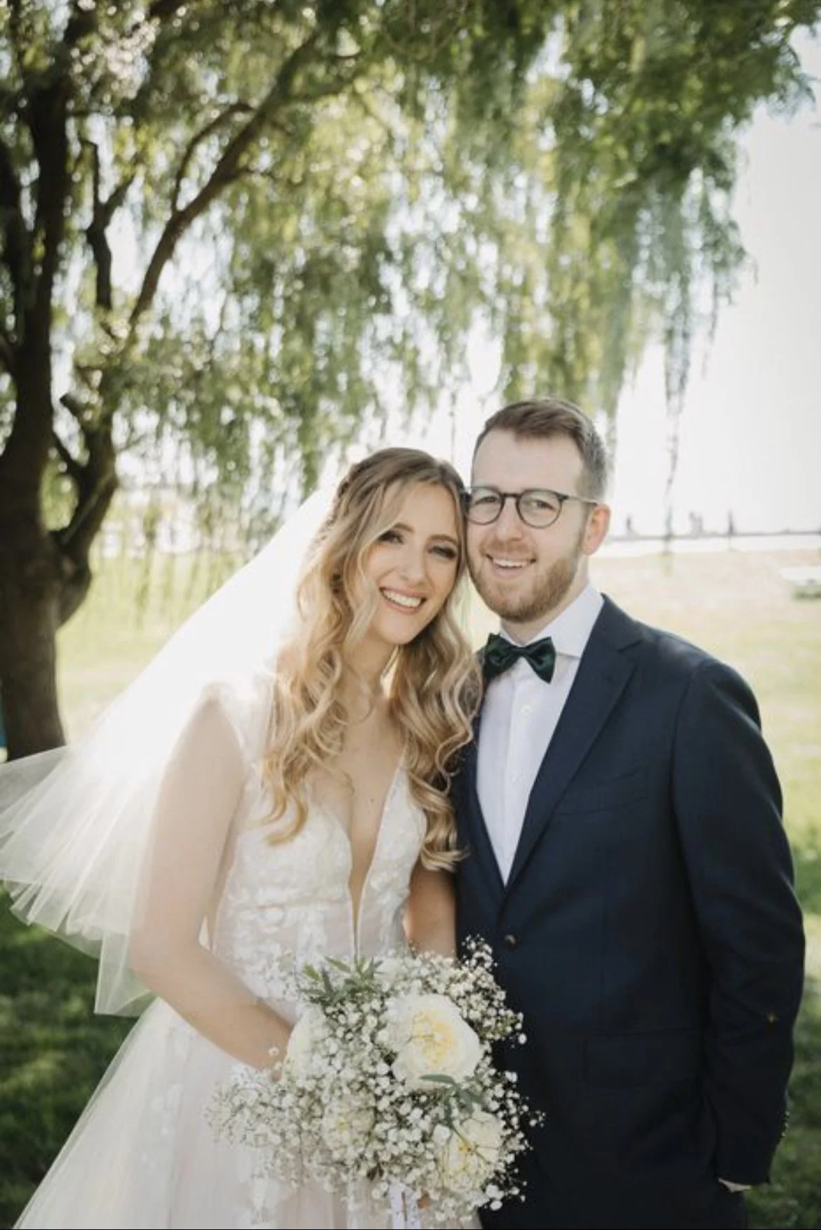 Bride and groom smiling at their outdoor wedding, standing under a tree. The bride holds a bouquet of white flowers.