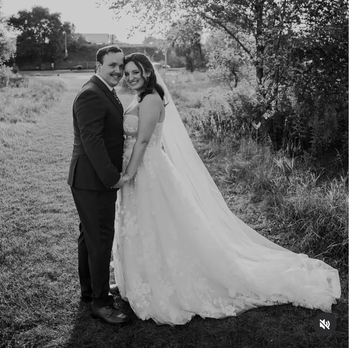 A black and white photo of a couple on their wedding day, standing outdoors on grass with trees in the background. The groom is wearing a suit, and the bride is in a wedding dress with a long veil.