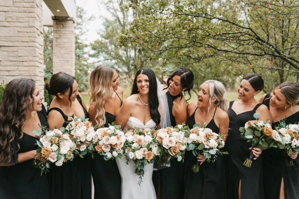 Bride in white dress surrounded by bridesmaids in black dresses, holding bouquets, smiling outdoors.
