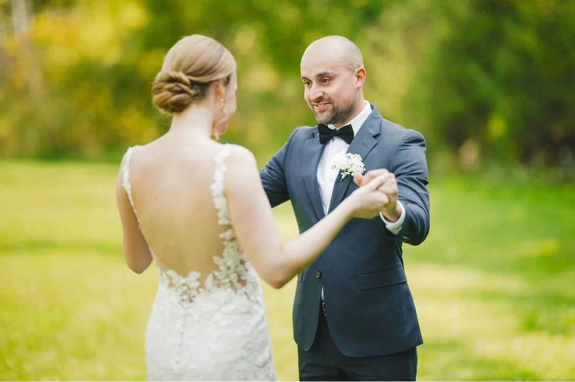 A groom in a gray suit dancing with a bride in a white wedding dress outdoors.