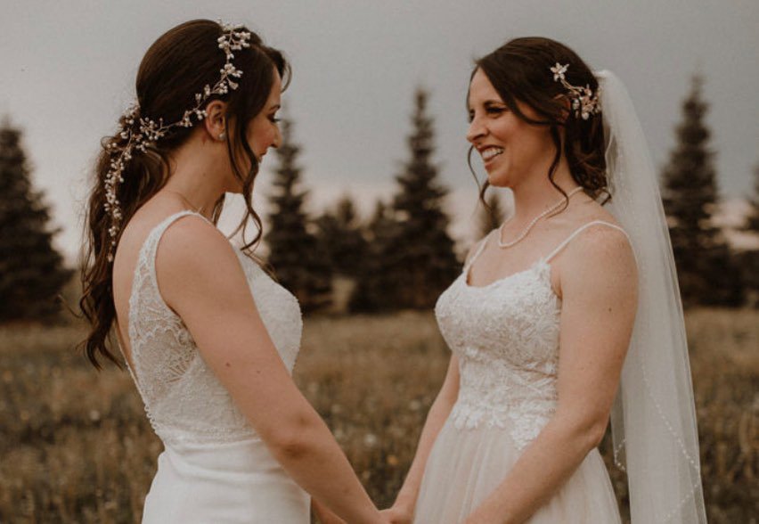 Two brides in white wedding dresses holding hands outdoors, smiling at each other with trees in the background.