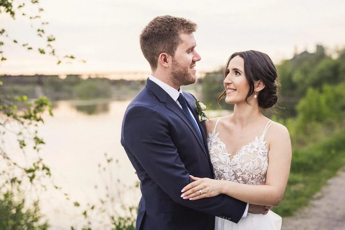 Bride and groom smiling at each other by a lakeside during sunset.