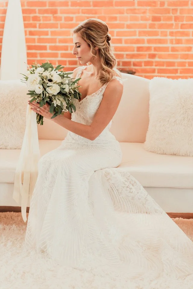 Bride sitting on a couch holding a bouquet, wearing a lace wedding dress, with a brick wall background.