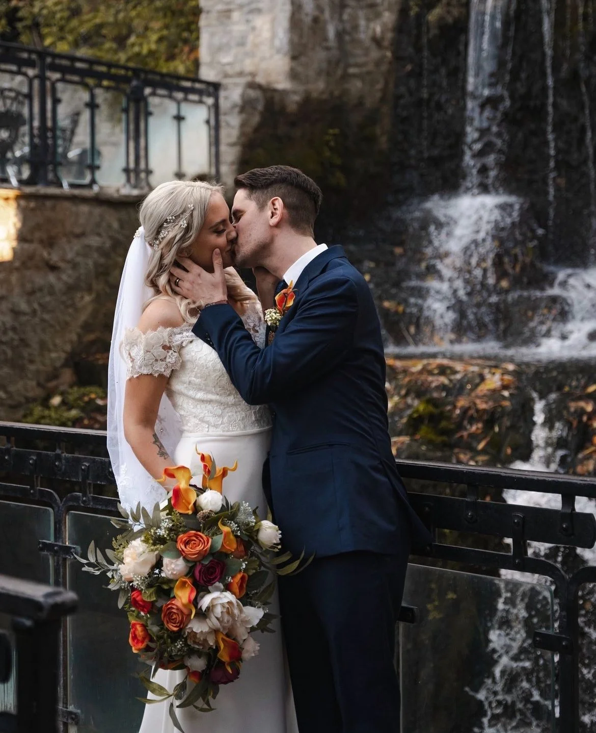 A bride and groom kissing in front of a waterfall, with the bride holding a bouquet of orange and white flowers. The groom is wearing a dark suit, and the bride is in a white dress with lace details.
