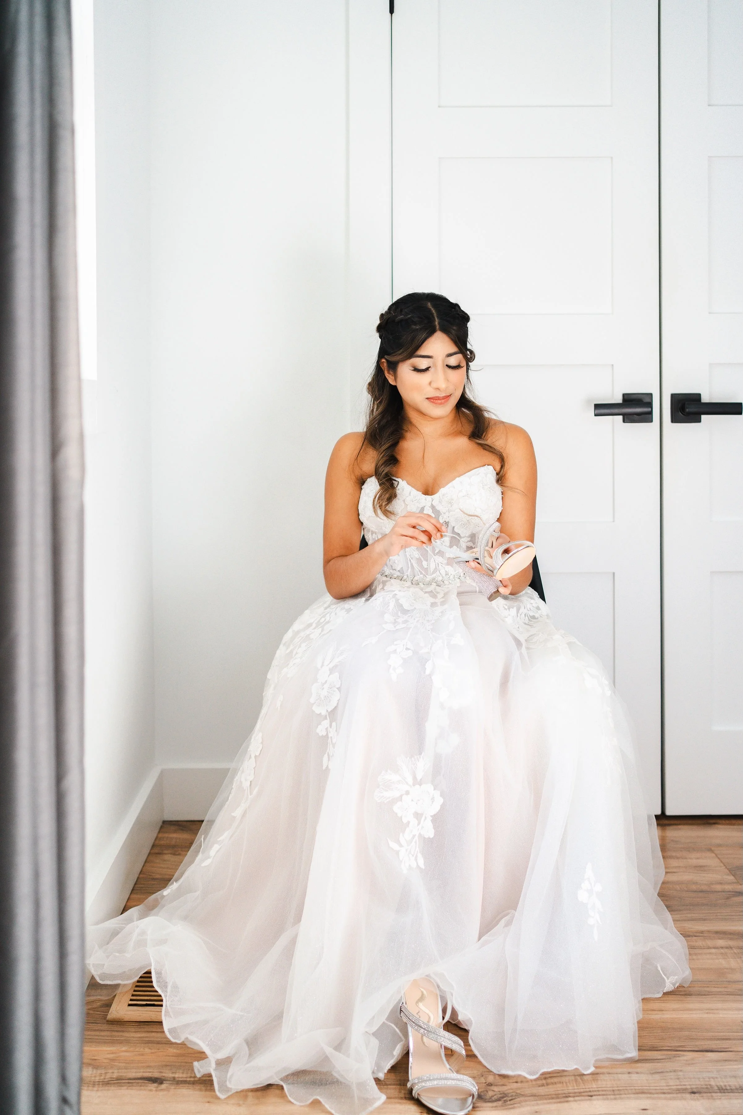 Bride in a white wedding dress sitting, holding makeup compact, near closed white doors.