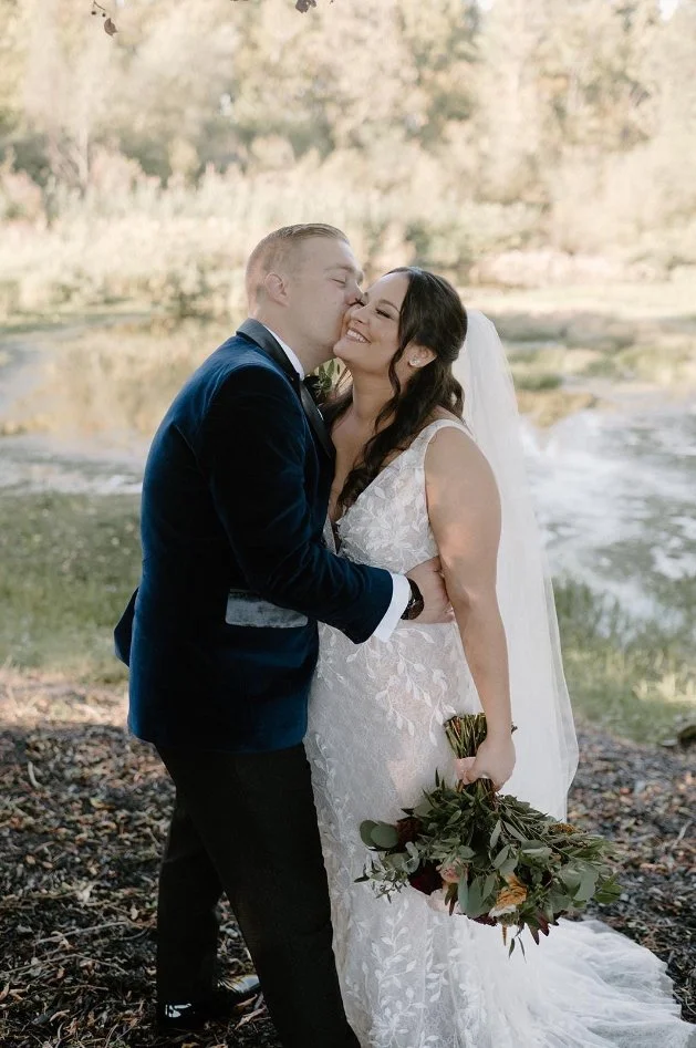 A couple in wedding attire embracing outdoors, with the groom kissing the bride's cheek and the bride holding a bouquet of flowers.
