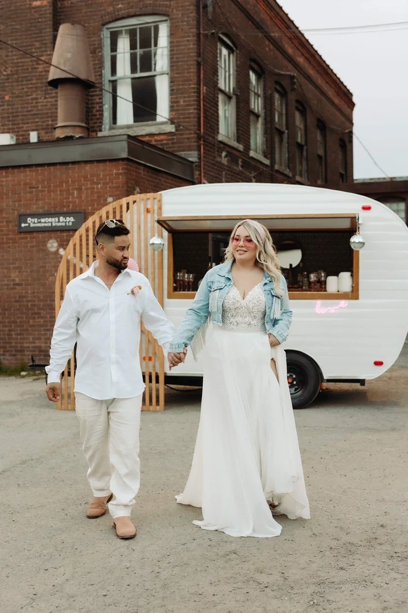 A couple holding hands in front of a white food truck, with a woman in a white dress and denim jacket, and a man in a white shirt and pants, standing outside a brick building.
