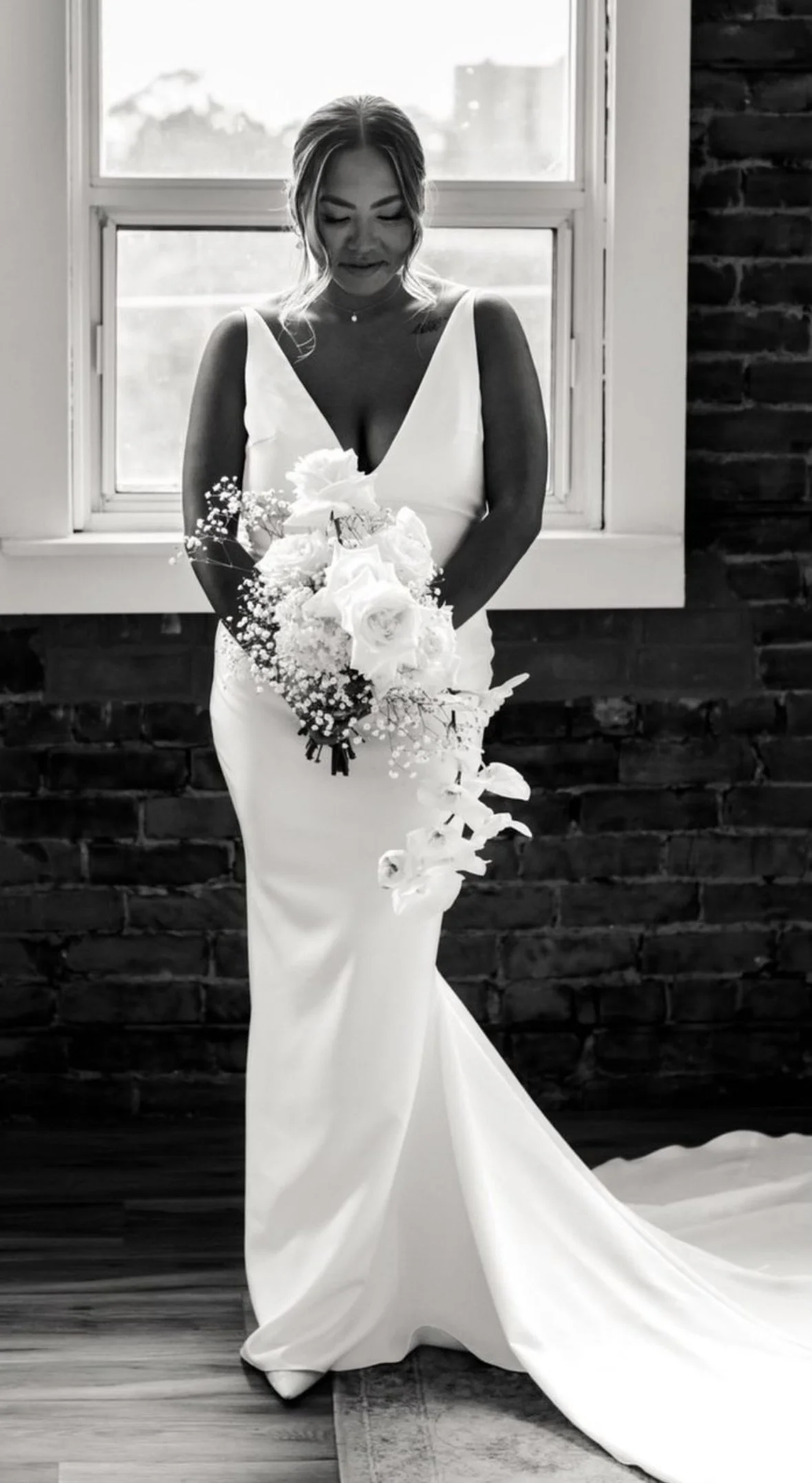 Black and white photo of a bride in a wedding dress holding a bouquet, standing indoors by a window.