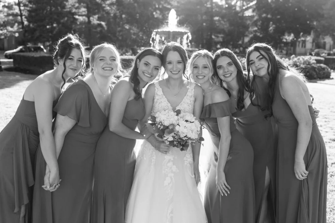 Black and white photo of a bride holding a bouquet, surrounded by bridesmaids in dresses, smiling in an outdoor setting with a fountain in the background.