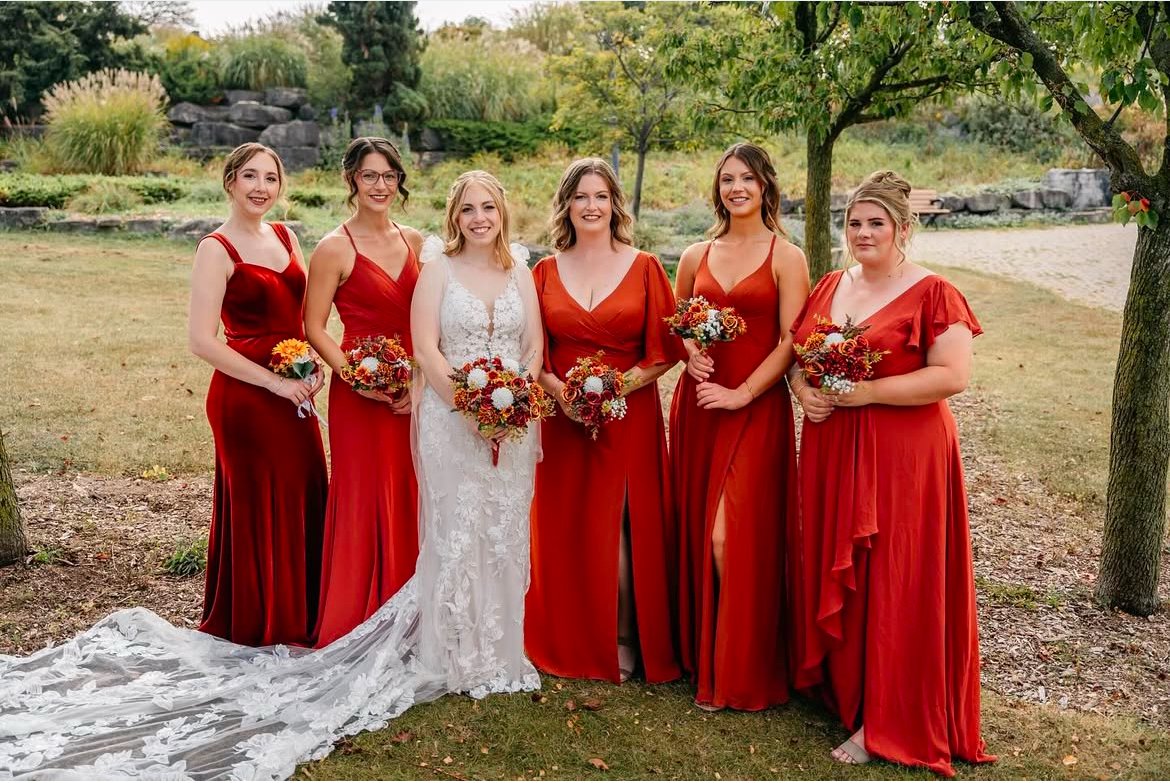 Bride with bridesmaids in red dresses holding bouquets outdoors.