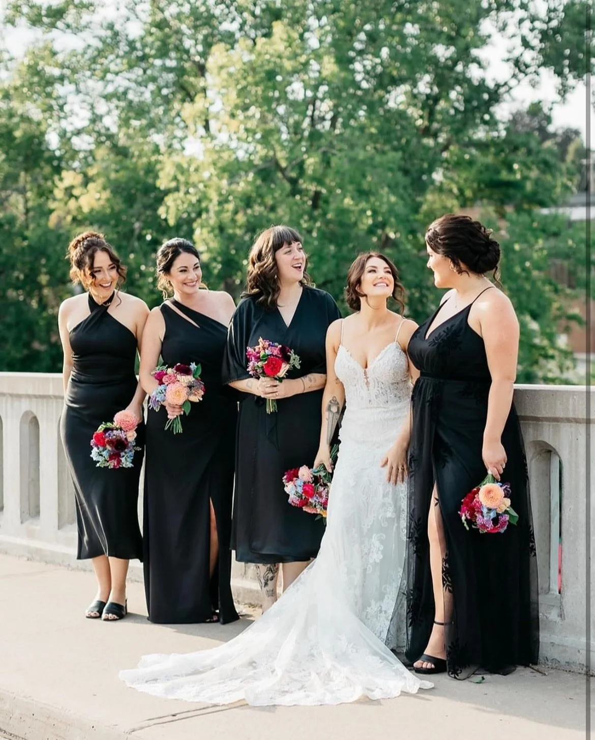 A bride in a white wedding dress stands on a bridge with four bridesmaids wearing black dresses, holding colorful bouquets, surrounded by greenery.