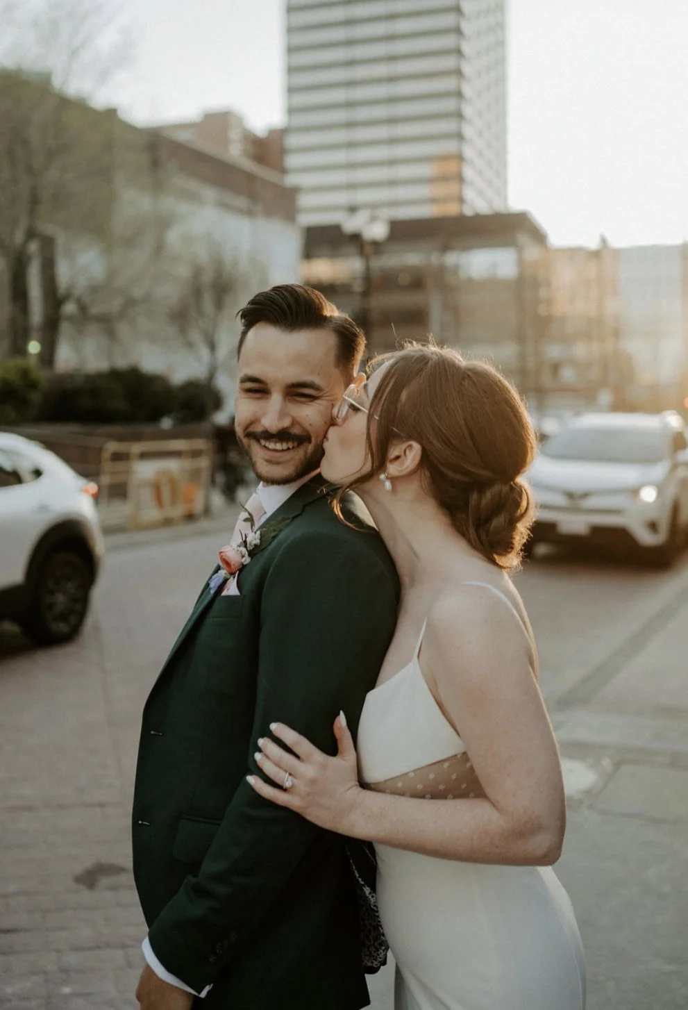 A couple dressed in formal attire, with a woman kissing a man on the cheek in a city setting during sunset.