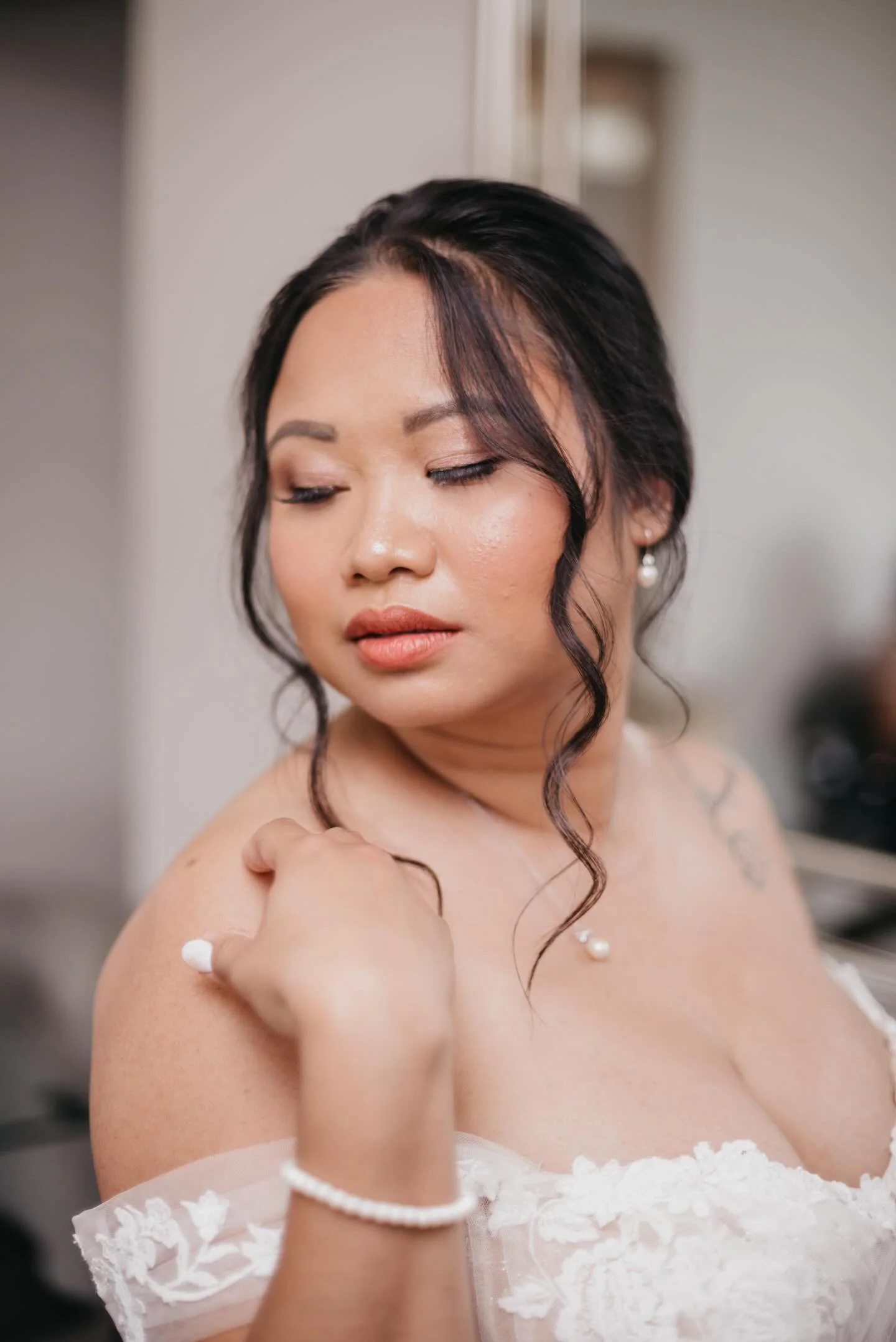 Woman in a wedding dress looking down, with hair styled in loose curls, wearing pearl jewelry.