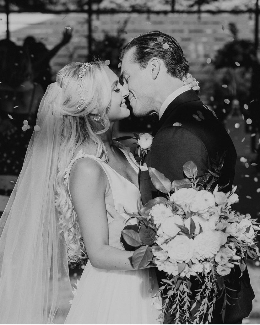Black and white photo of a newlywed couple kissing, bride holding a bouquet of flowers.