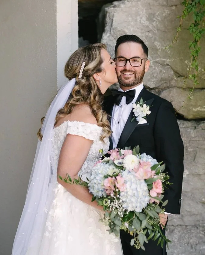 Bride kissing groom on cheek, both smiling, holding a floral bouquet, wearing wedding attire.