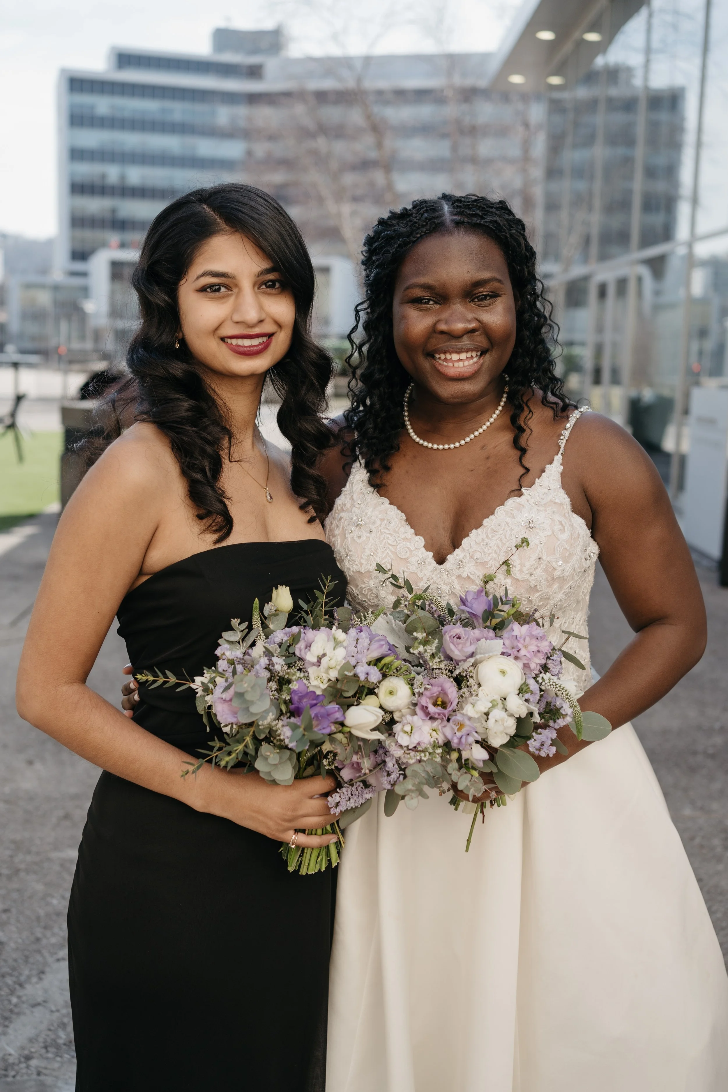 Two women smiling, one in a wedding dress holding a bouquet, outdoors with a city building in the background.
