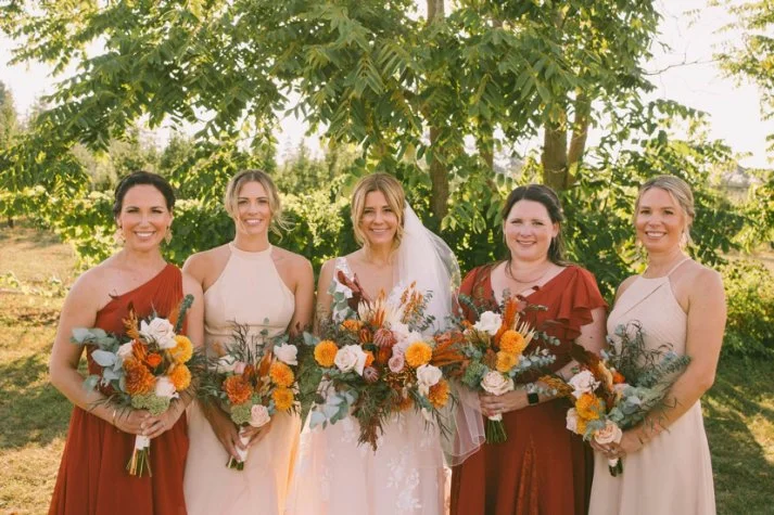 Bride and bridesmaids in a garden, holding colorful bouquets, wearing a mix of white and terracotta dresses.
