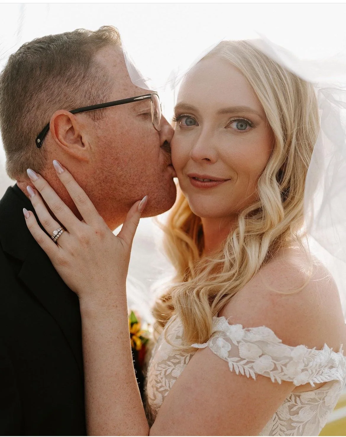 Bride and groom, the groom kissing the bride on the cheek, bride wearing an off-shoulder lace gown and veil, close-up portrait, smiling.