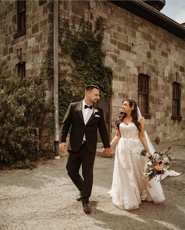 Bride and groom walking hand in hand outside a stone building, with greenery in the background.