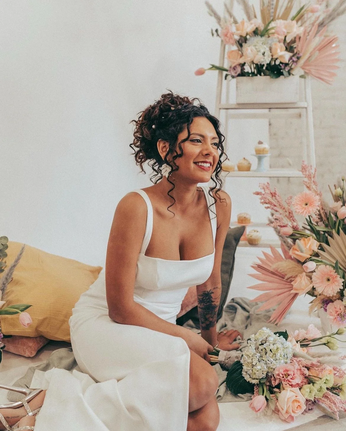 Smiling woman with curly hair in white dress sitting beside floral arrangements and a ladder with flowers and cupcakes.