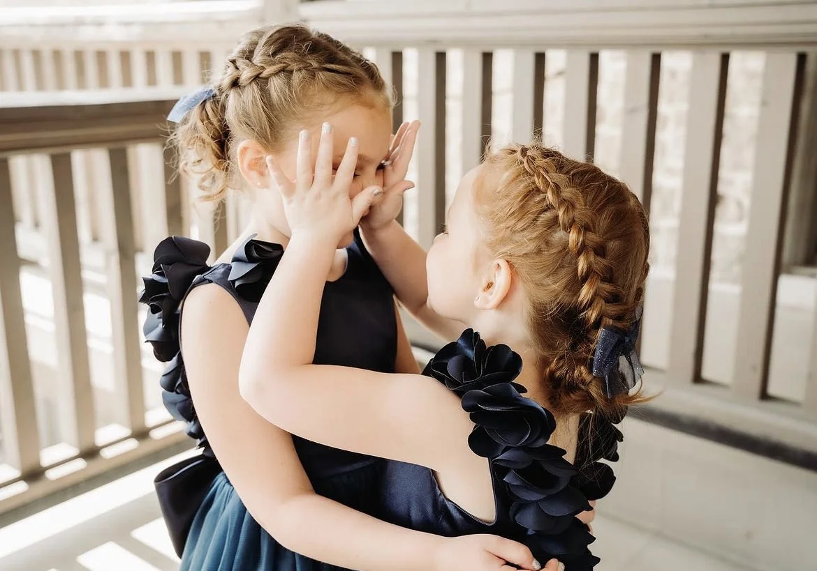 Two young girls in navy blue dresses with braided hair touch each other's faces playfully.