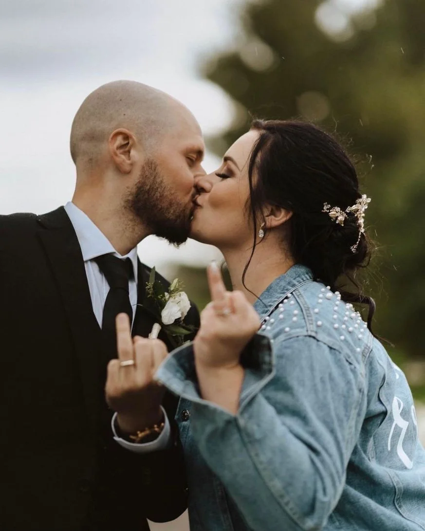 A couple kissing, with the woman wearing a denim jacket and both showing raised middle fingers. The man is in a suit with a boutonnière.