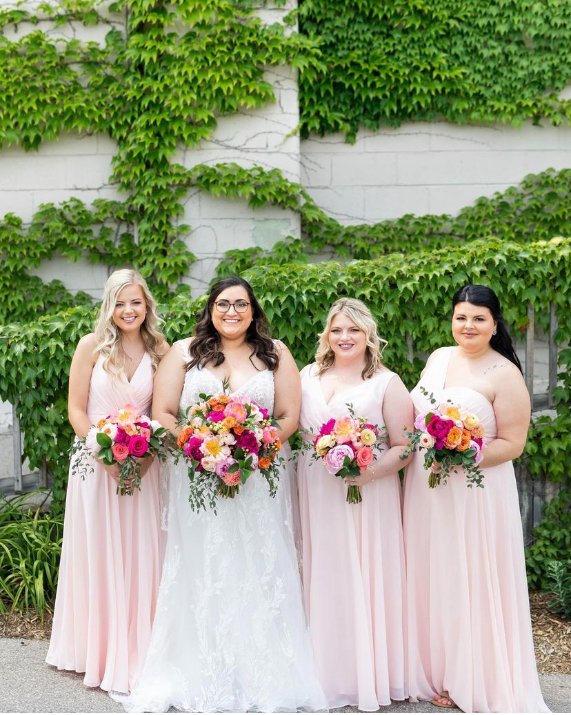 Bride and bridesmaids in pink dresses holding colorful flower bouquets, standing in front of a wall with green ivy.