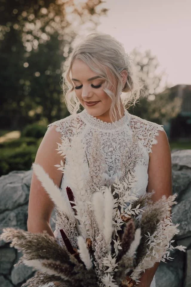 A bride in a lace wedding dress holding a bouquet of dried flowers outdoors.
