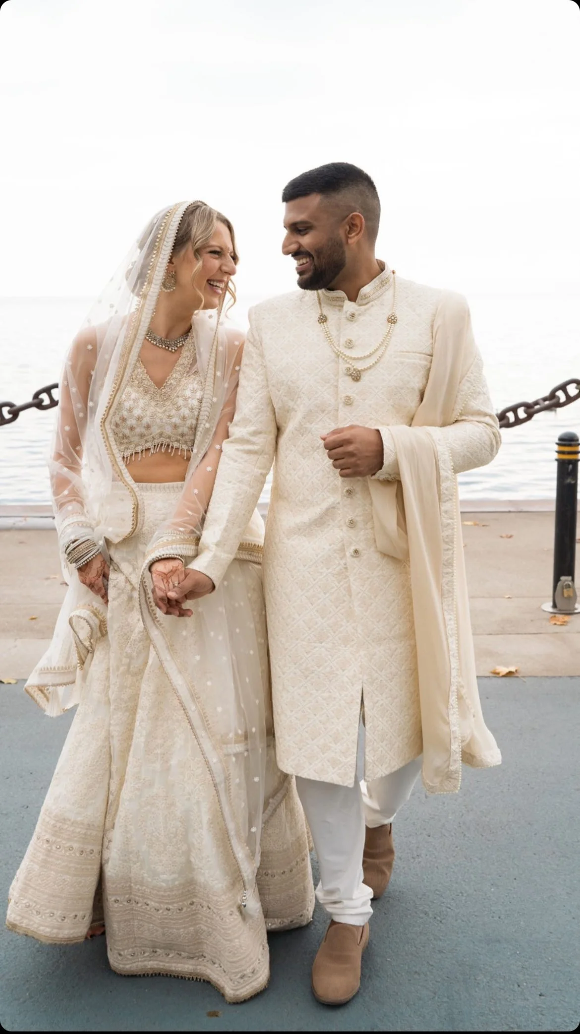 A couple in traditional Indian wedding attire, holding hands and smiling at each other near a waterfront. The bride is wearing an ornate white lehenga with a sheer dupatta, and the groom is in a white sherwani with accessories.