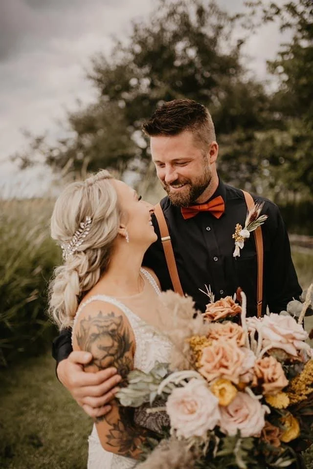 A couple in wedding attire smiling at each other outdoors, surrounded by greenery. The woman holds a large bouquet of flowers, and has a decorative hairpiece. The man is wearing a black shirt with suspenders and a bow tie.