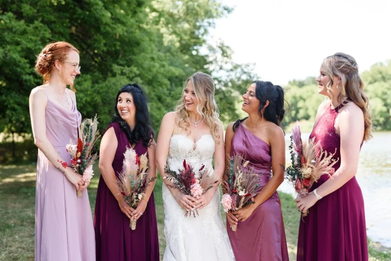 Bride and four bridesmaids in various shades of purple dresses, holding bouquets, standing outdoors with trees and water in the background, smiling and talking.