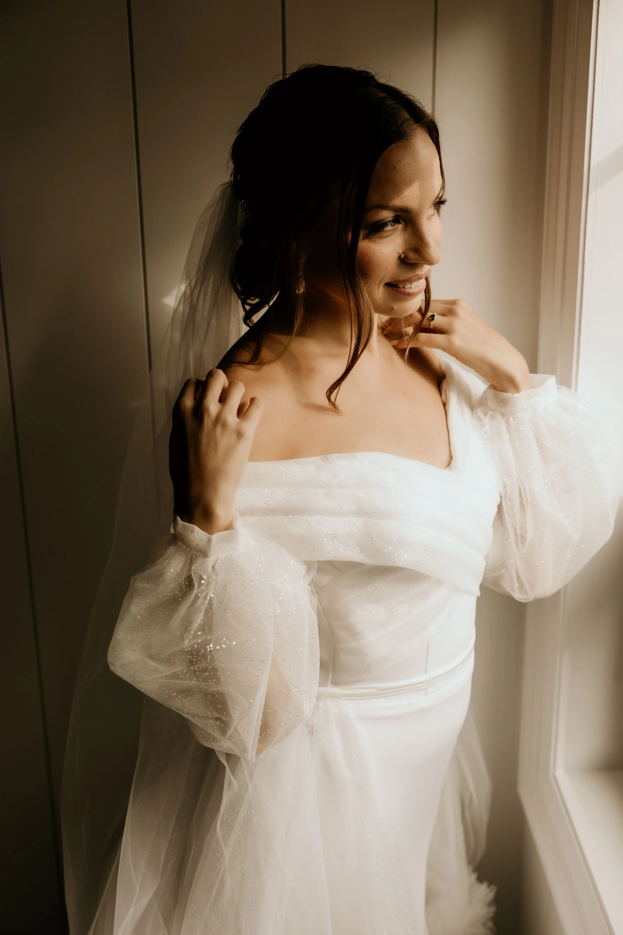 Bride in white wedding dress with sheer sleeves and veil, looking out a window.