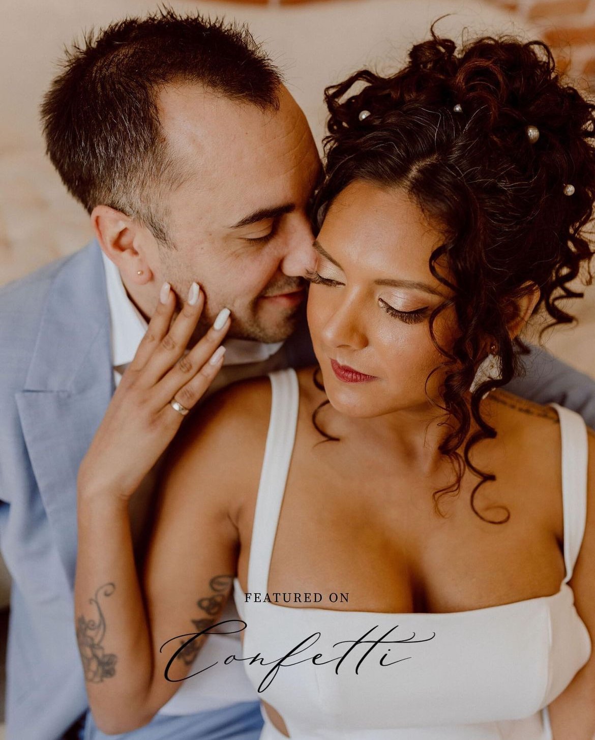 A couple embracing closely, the man wearing a light blue suit and the woman in a white dress with curly hair adorned with beads. Text over the image reads "Featured on Confetti."
