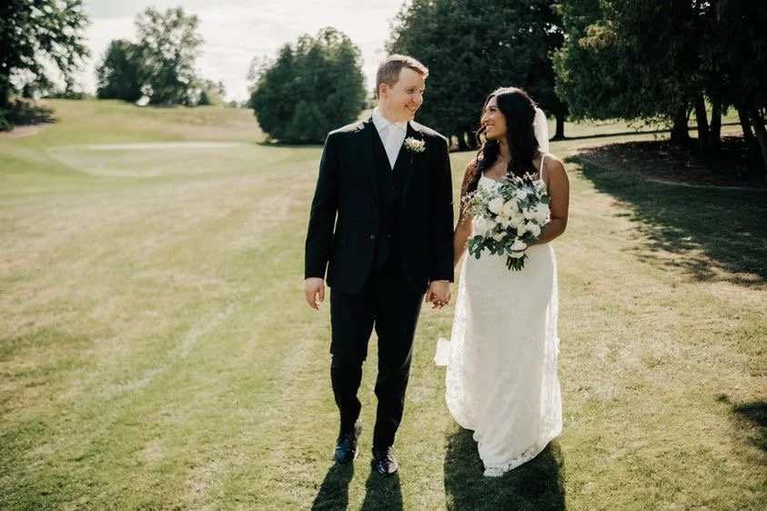 A couple walking hand in hand on a grassy field, with the woman in a white wedding dress holding a bouquet, and the man in a black suit, smiling at each other.