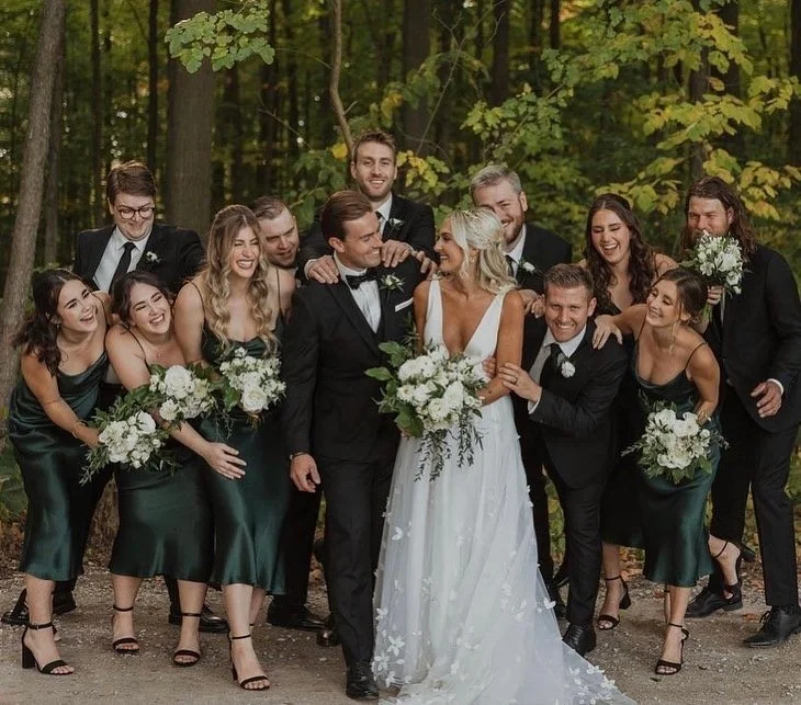 Wedding party in a forest setting with bride, groom, bridesmaids, and groomsmen celebrating and smiling.