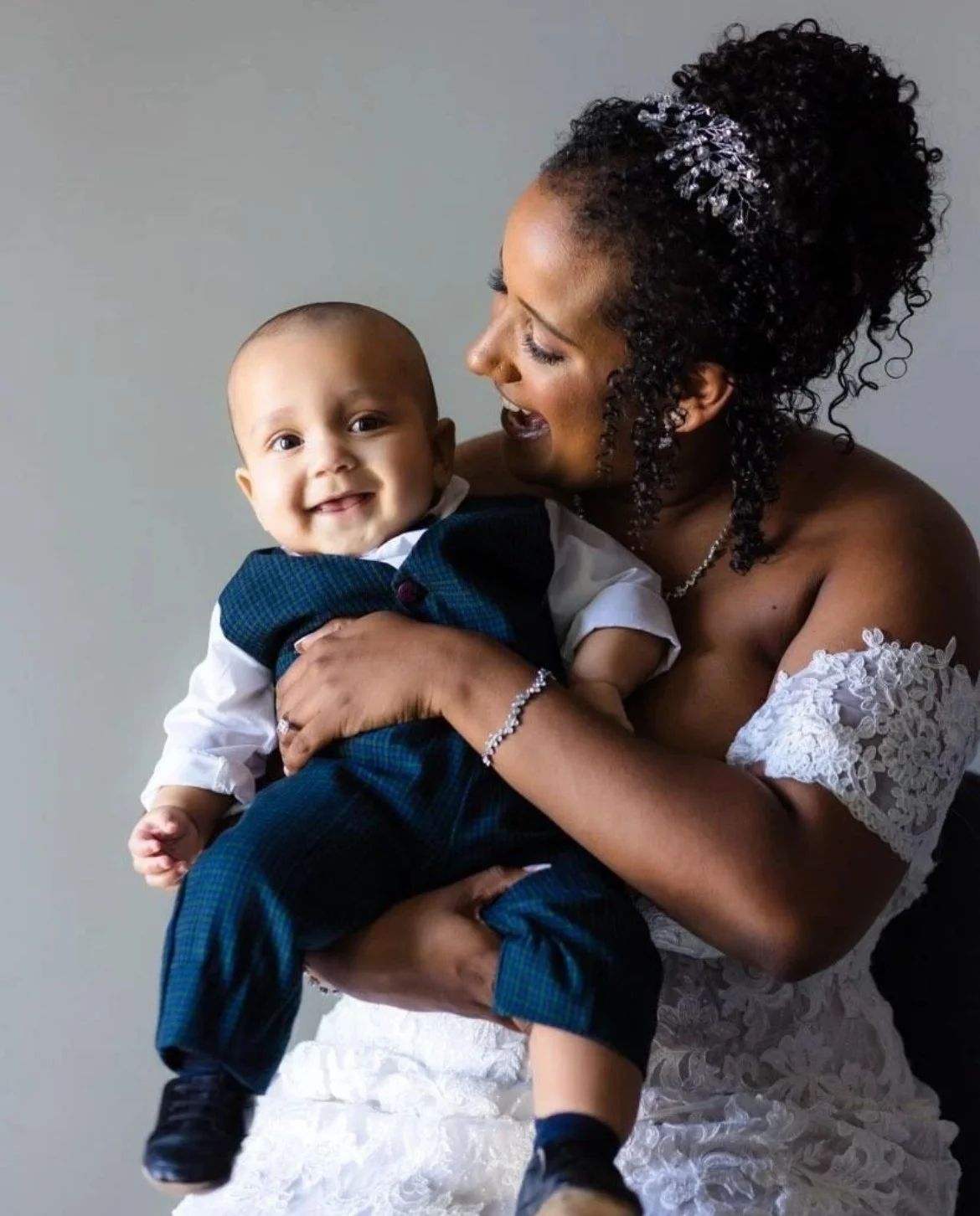 A woman in a white lace dress holding a smiling baby dressed in a dark suit. The background is neutral.