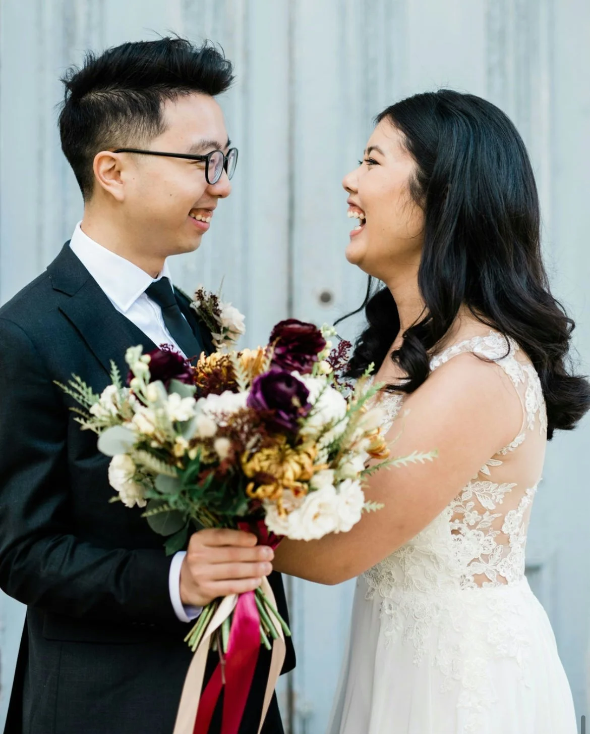 Bride and groom smiling while holding a bouquet of flowers, bride in a white lace dress, groom in a dark suit and glasses.